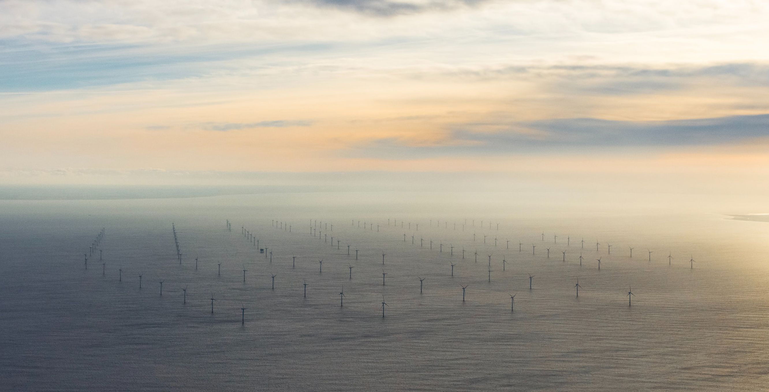 An aerial view shows a large offshore wind farm with numerous wind turbines standing in the sea. The sky is partly cloudy, and the sun is shining through, creating soft lighting.