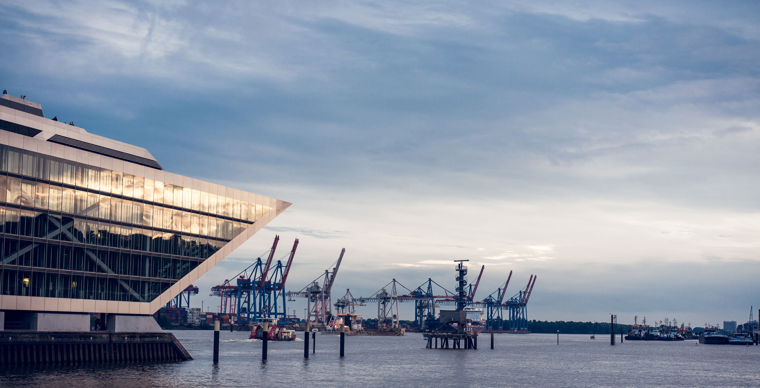 Ein modernes Gebäude mit spitzer Fassade befindet sich am Wasser. Im Hintergrund sind mehrere Kräne im Hafen zu sehen. Der Himmel ist bewölkt und das Wasser reflektiert das Licht der Umgebung.