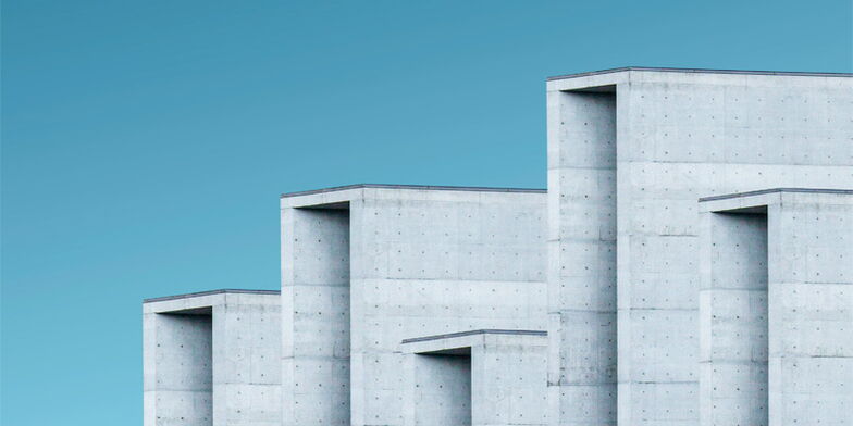 Graue Betongebäude mit minimalistischen, rechteckigen Formen erstrecken sich stufenartig gegen einen klaren blauen Himmel. Die Oberflächen sind glatt und modern gestaltet.