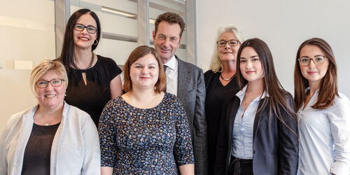 Eine Gruppe von sieben Personen, vier Frauen vorne und drei Personen, darunter zwei Frauen und ein Mann, hinten, steht in einem Büro vor einer Wand. Alle lächeln in die Kamera.