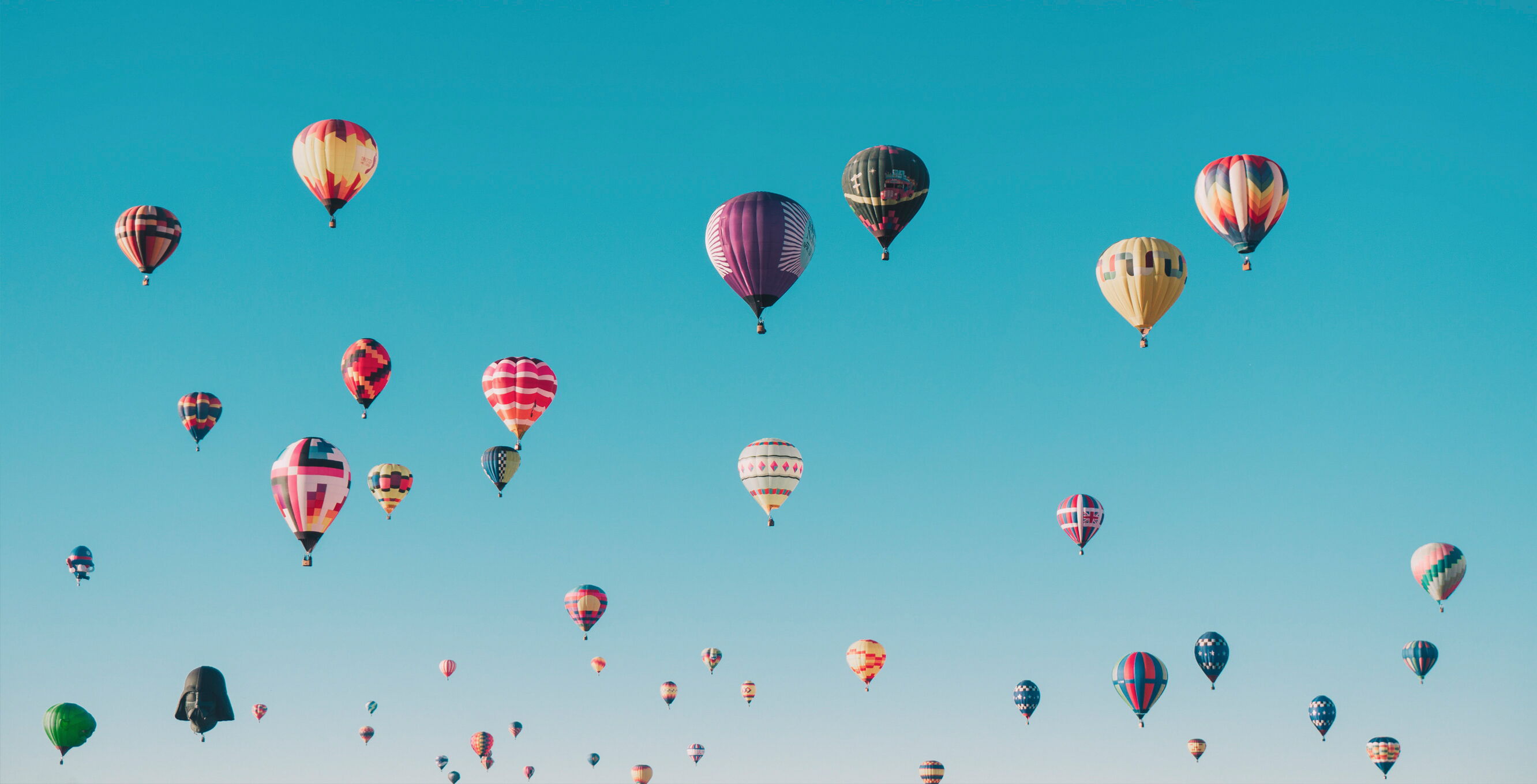 Several colorful hot-air balloons in various designs float in the sky against a clear blue background.
