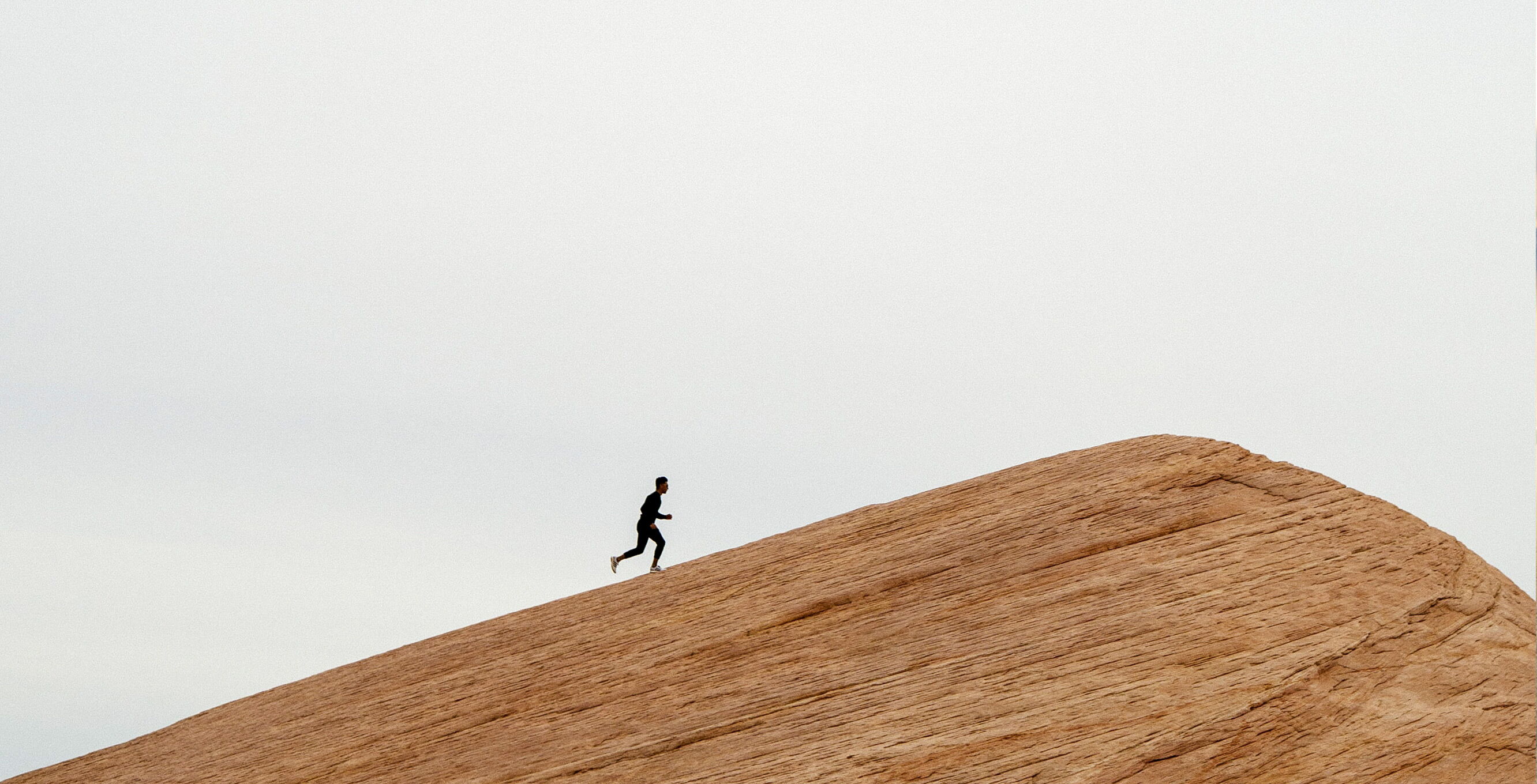 Eine Person läuft einen felsigen, schrägen Hügel hinauf. Der Himmel ist bewölkt. Die Szene wirkt minimalistisch und die Person ist als Silhouette zu sehen.