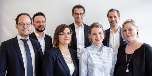 A group of seven people in business attire is standing in front of a white wall. Four men and three women are smiling at the camera. Everyone is wearing formal clothing, such as suits and blouses.